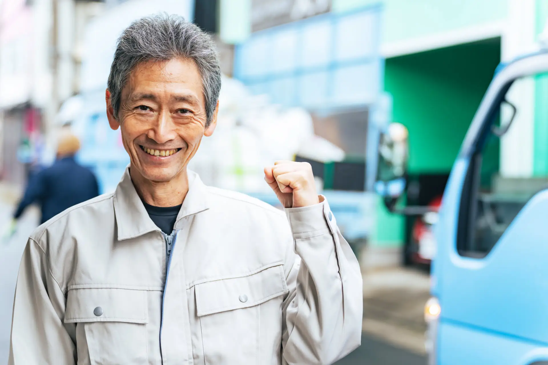 青いトラックの前で、笑顔でガッツポーズをする作業服姿の男性